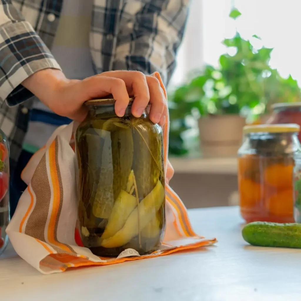 A person placing a lid on a large jar of homemade pickles, using a kitchen towel for grip. The jar contains whole pickles submerged in brine, with other jars and fresh cucumbers visible on the table. The image aligns with the title "Are Pickles Good for Weight Loss? Truth Explained.