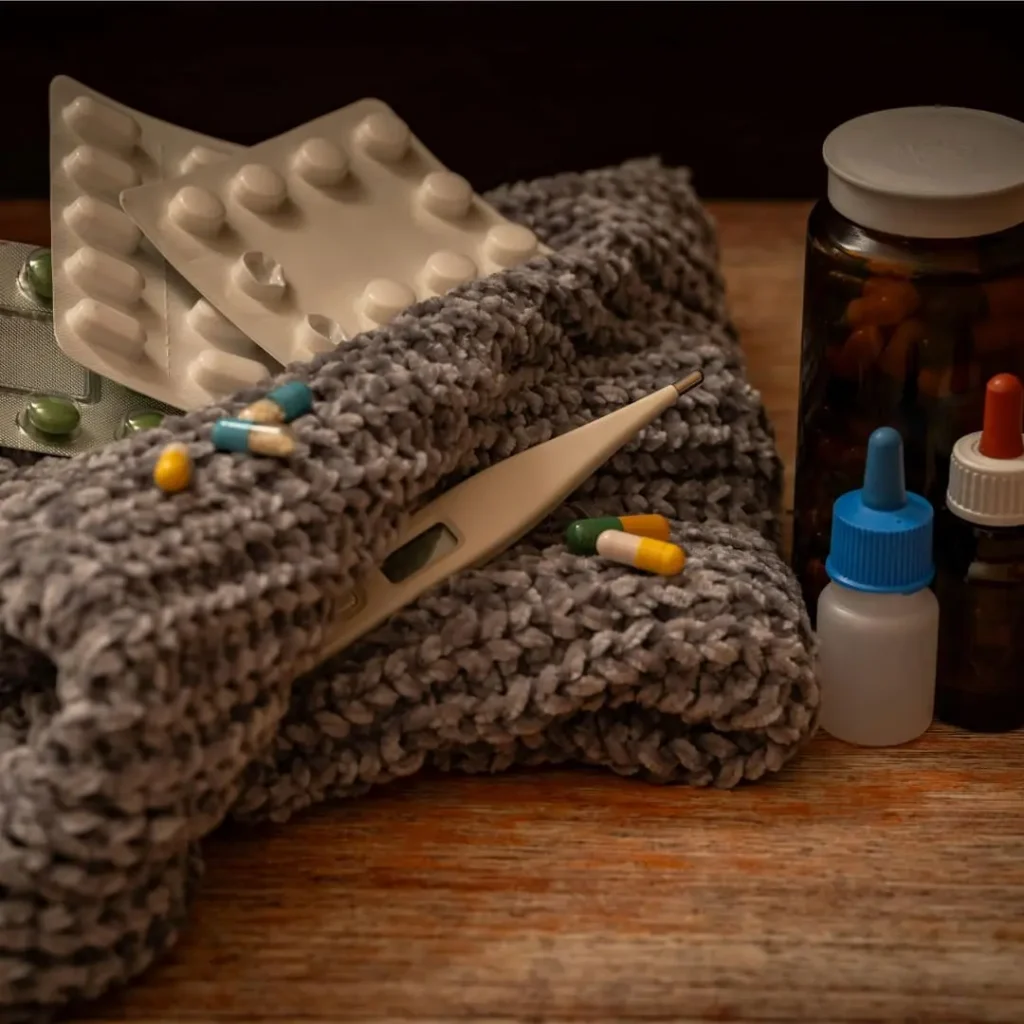 A collection of medical supplies, including blister packs of pills, a thermometer, a knitted scarf, and bottles of medicine, displayed on a wooden surface.