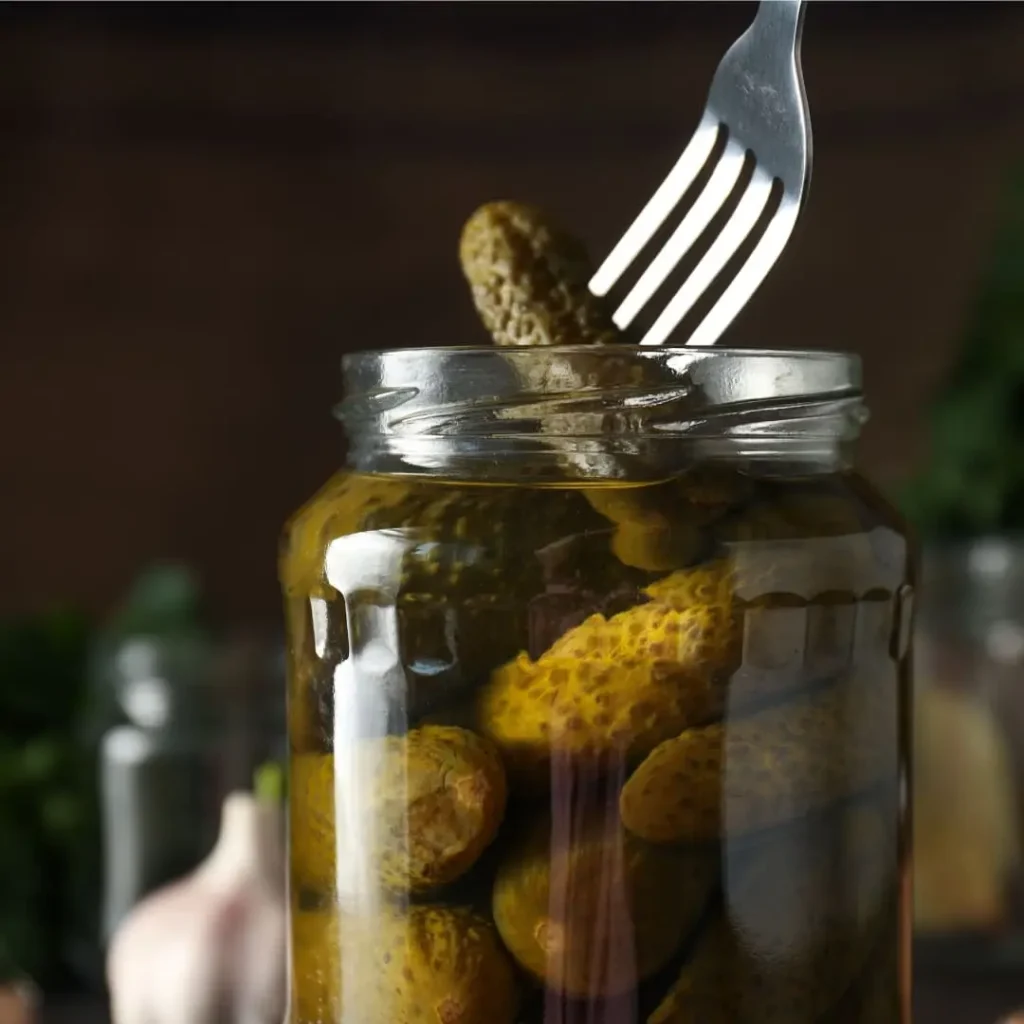 A jar of pickles with a fork pulling out one of the pickles. The jar is filled with whole pickles, submerged in brine, with a rustic kitchen setting in the background.