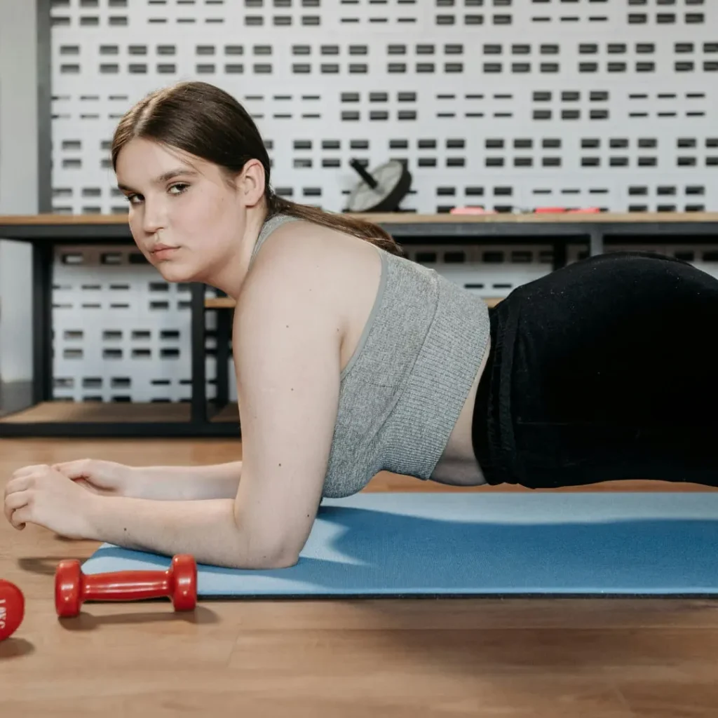 A woman in a gray sports bra and black leggings performing a plank on a blue exercise mat in a gym setting, with dumbbells placed nearby.