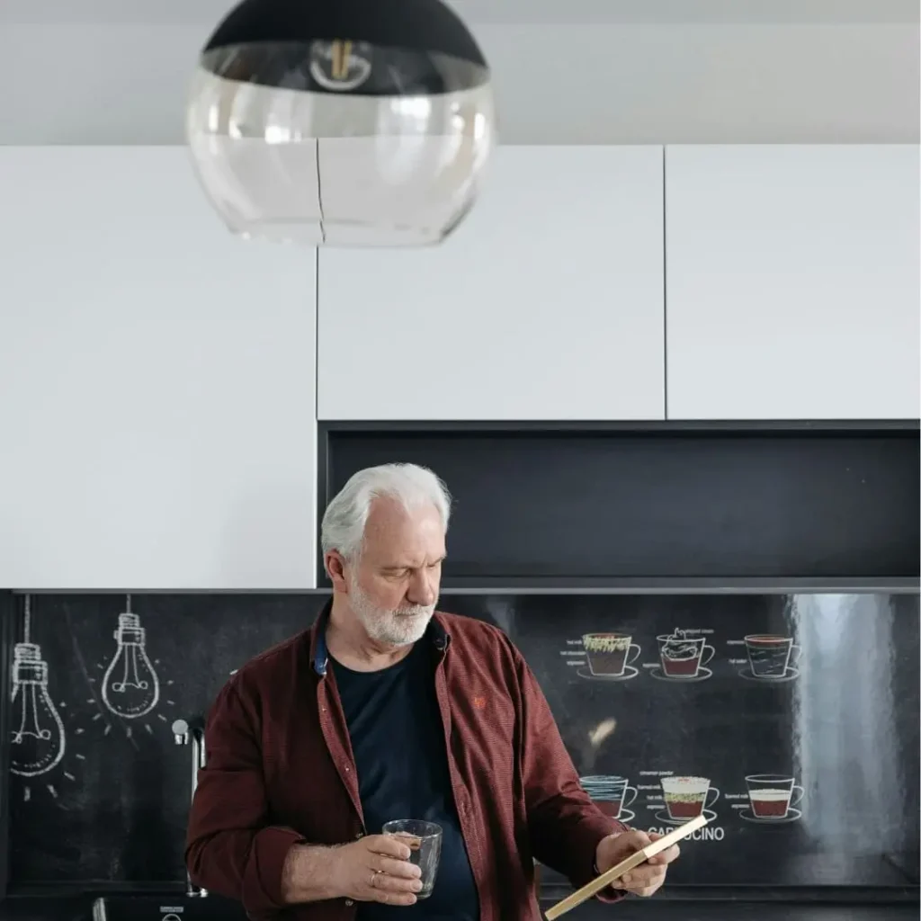 An older man with white hair and a beard standing in a modern kitchen, holding a glass and reading from a wooden board, with a coffee-themed chalkboard in the background.