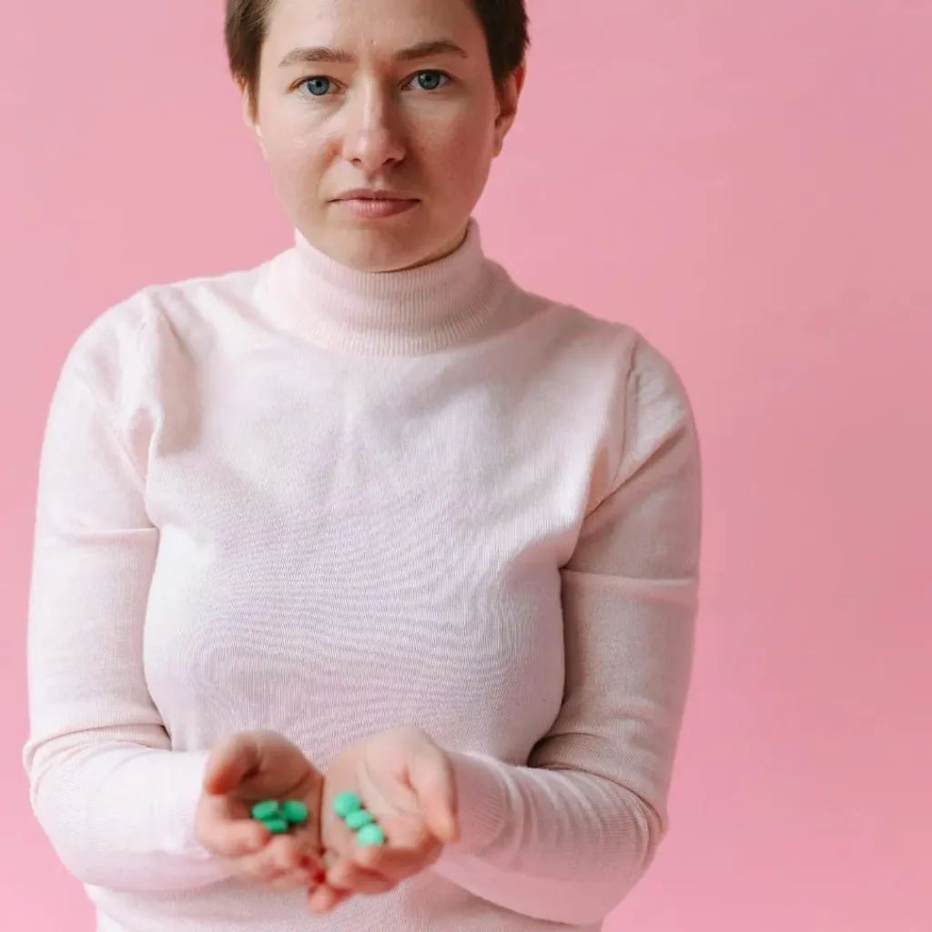 A woman in a light pink turtleneck sweater standing against a pink background, holding green pills in her open hands with a serious expression.