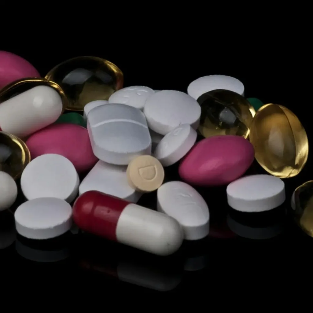 A close-up view of assorted colorful pills and capsules, including white, pink, gold, and red medication, displayed on a black surface.