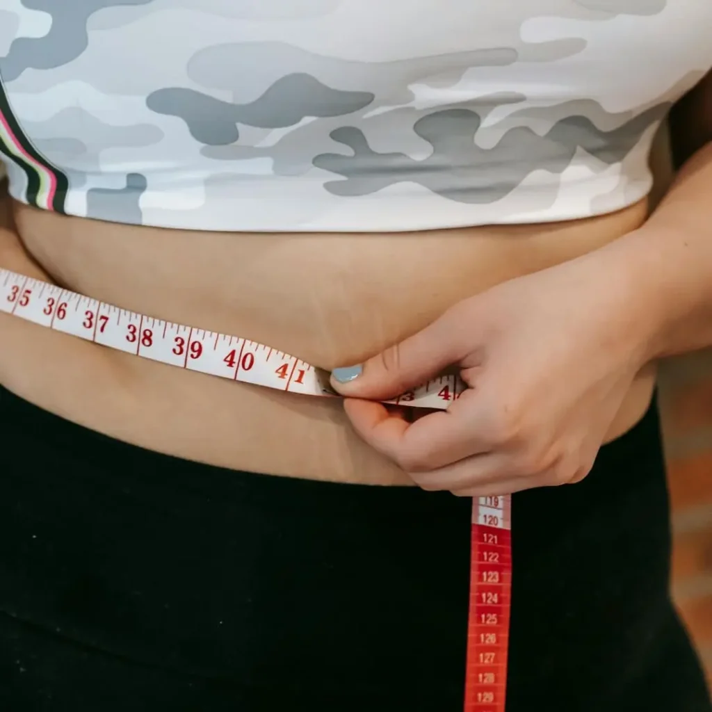 A person measuring their waist with a tape measure while wearing a camouflage-patterned crop top and black leggings.