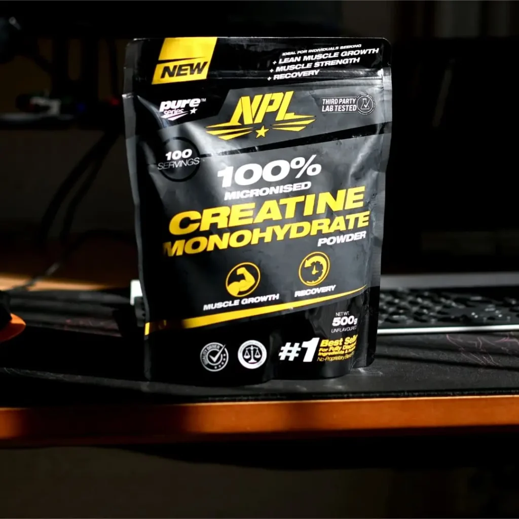 A black and yellow pouch of NPL 100% Micronised Creatine Monohydrate Powder placed on a desk with a gaming setup in the background.