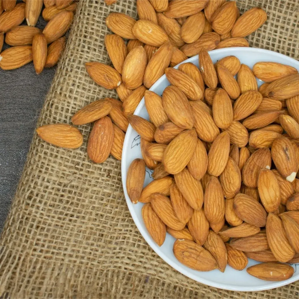 A white bowl filled with raw almonds spilling onto a textured burlap mat, showcasing their natural brown color and texture.