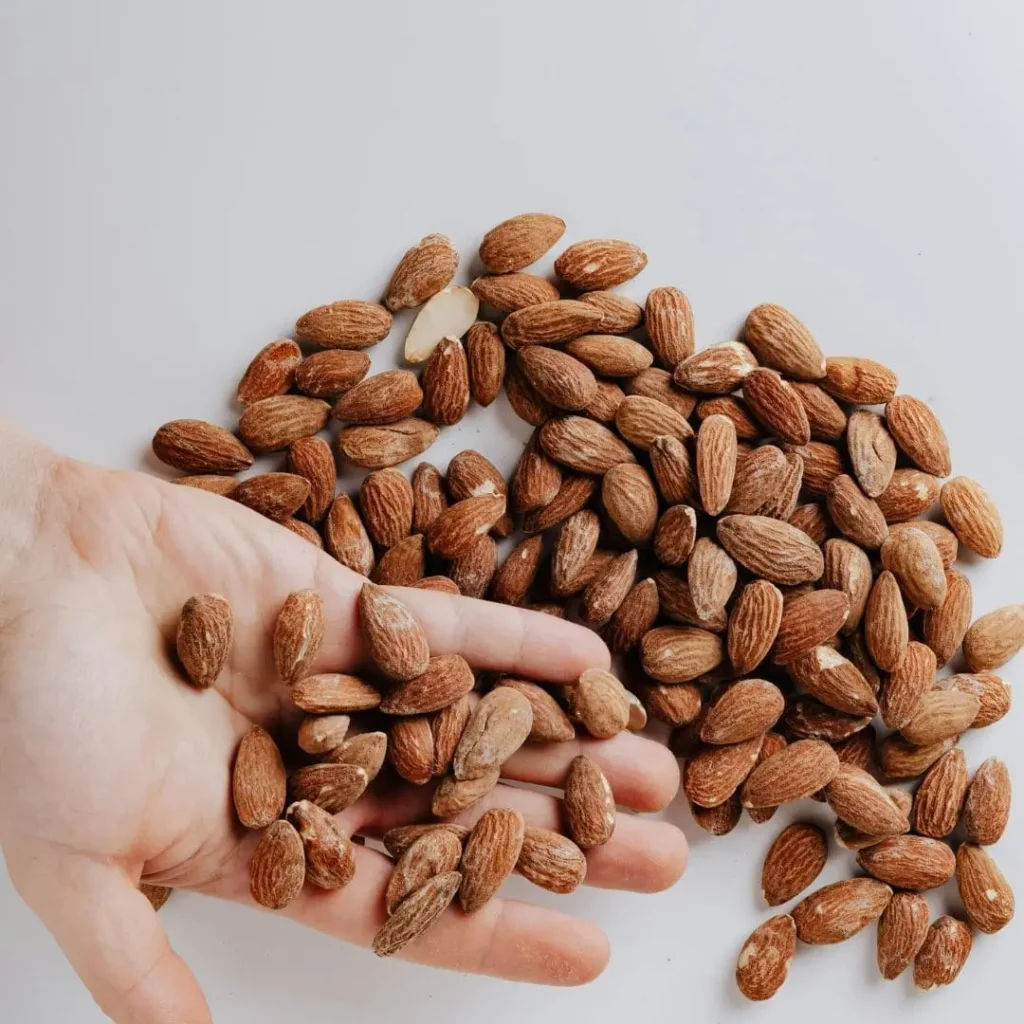 A close-up of a hand holding raw almonds with more almonds scattered on a clean white surface, highlighting their natural texture and earthy tones.