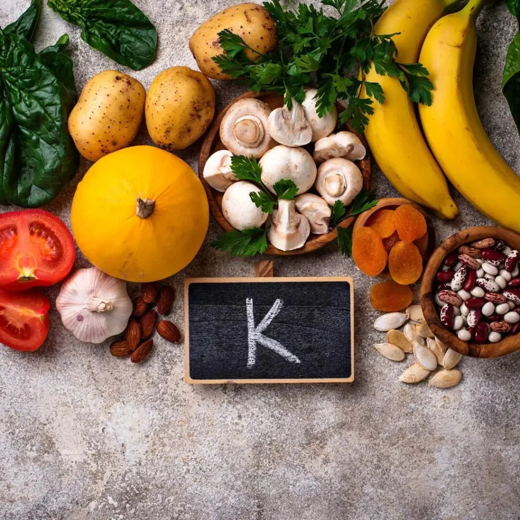 An assortment of potassium-rich whole foods, such as bananas, potatoes, mushrooms, spinach, tomatoes, dried fruits, nuts, and legumes, arranged with a small chalkboard sign displaying the letter "K," the chemical symbol for Potassium Supplements.