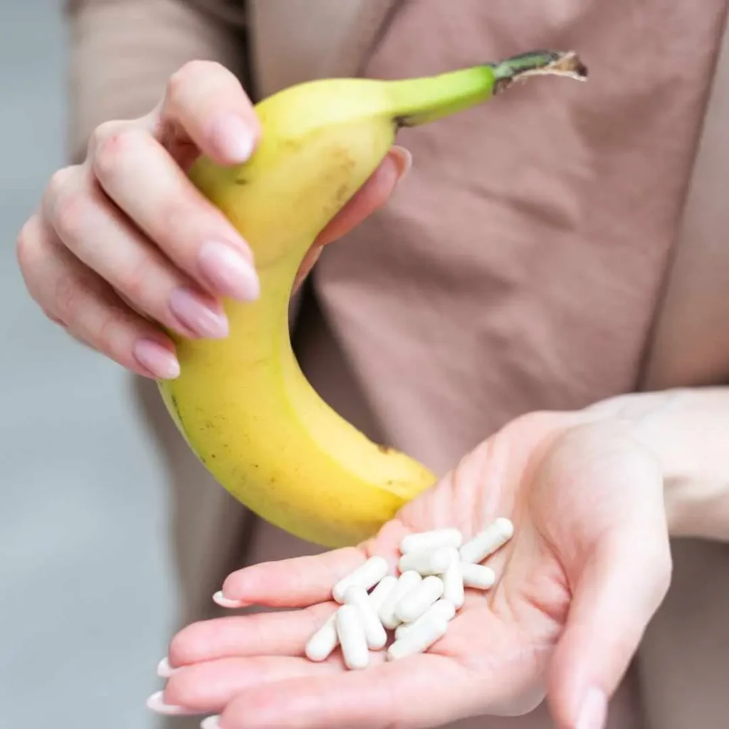 A person holding a fresh banana in one hand and a handful of white potassium supplement capsules in the other, comparing natural vs. supplemental sources of potassium.