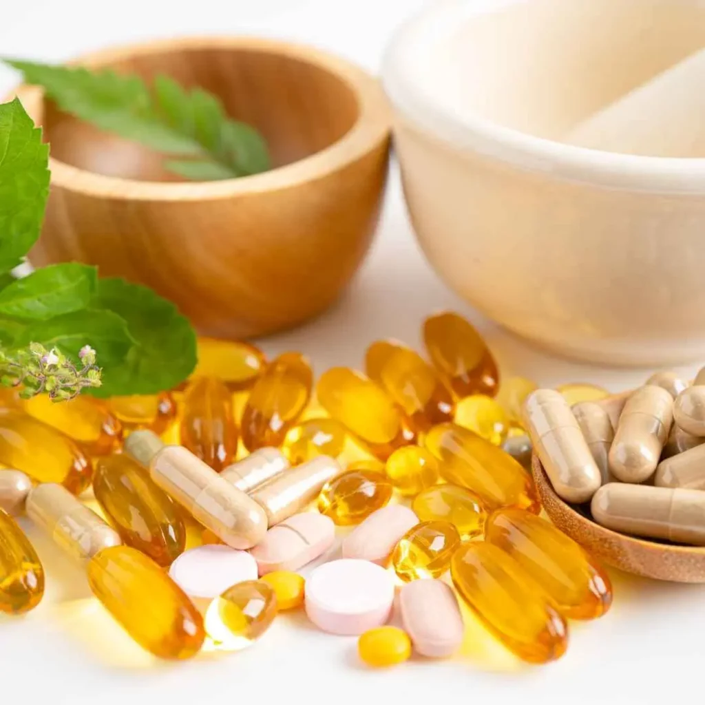 A collection of various dietary supplements, including soft gel capsules, tablets, and herbal capsules, placed near wooden and ceramic bowls with fresh green leaves.