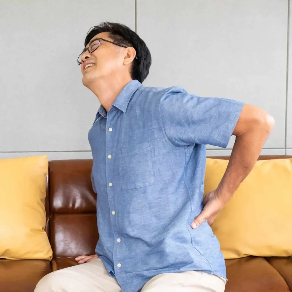 An older man wearing a blue shirt, sitting on a couch and clutching his lower back in discomfort, possibly indicating symptoms of potassium imbalance or deficiency-related cramps.